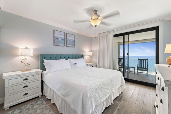 Ein Schlafzimmer mit einem Bett, Nachttischen, einem Deckenventilator, Meerblick durch eine Schiebetür und einem Balkon mit Stühlen.