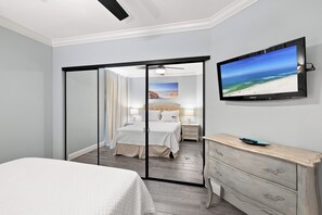 A hotel room with a bed, a television, a dresser, and a view of the beach through a glass door. - Windward Pointe 1204 by Liquid Life (Orange Beach)