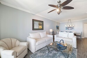 A living room with a ceiling fan, a white sofa, a beige armchair, and a glass coffee table with decorative items. - Windward Pointe 1204 by Liquid Life (Orange Beach)