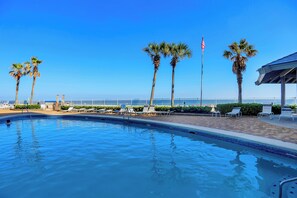 A swimming pool with lounge chairs and palm trees, a flag, and a view of the ocean. - Windward Pointe 1204 by Liquid Life (Orange Beach)