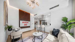 Ein modernes Wohnzimmer mit einem 'BOOK ME' Schild, einem Essbereich, einer Küche und einer großen Küstpflanze.