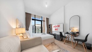 A modern hotel room with a bed, a desk, a chair, a sofa, a large mirror, and a 'BOOK ME' sign. - Free Housekeeping Visits During Your Stay - Stayshort - Luxury Studio Apartment in Meydan With Balcony (Dubai)