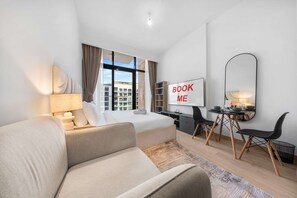 A modern hotel room with a bed, a desk, a chair, a sofa, a large mirror, and a 'BOOK ME' sign. - Free Housekeeping Visits During Your Stay - Stayshort - Luxury Studio Apartment in Meydan With Balcony (Dubai)