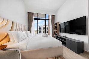 A modern hotel room with a large bed, a flat-screen TV, a wooden side table, and a view of the cityscape. - Free Housekeeping Visits During Your Stay - Stayshort - Luxury Studio Apartment in Meydan With Balcony (Dubai)