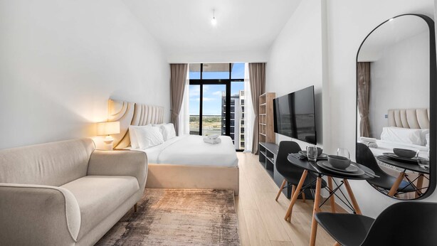 A modern hotel room with a bed, a sofa, a dining table with chairs, a TV, and a large window offering a city view. - Free Housekeeping Visits During Your Stay - Stayshort - Luxury Studio Apartment in Meydan With Balcony (Dubai)