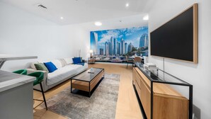A modern living room with a cityscape mural, a sofa, a coffee table, a TV, and a desk area. - Free Housekeeping Visits During Your Stay - Stayshort - Comfortable 1 Bedroom in Marina With Balcony (Dubai)