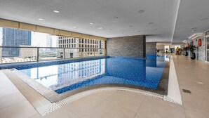 An indoor swimming pool with clear blue tiles, a tiled floor, and a view of a cityscape through large windows. - Free Housekeeping Visits During Your Stay - Stayshort - Comfortable 1 Bedroom in Marina With Balcony (Dubai)