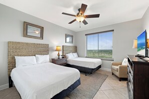 Ein Hotelzimmer mit zwei Betten, einem Deckenventilator, einem großen Fenster mit Jalousien, einem Fernseher und einem Schreibtisch mit Lampe.