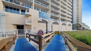 L’entrée d’un hôtel, avec un tapis bleu menant à une terrasse en bois, un panneau indiquant l’emplacement des douches et des points d’eau, et un bâtiment doté de plusieurs balcons.