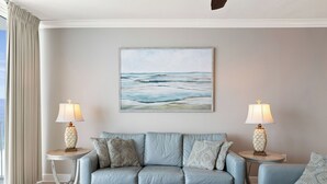 Un salon avec un canapé bleu clair, deux tables d’appoint avec des lampes, une table basse en osier et un tableau représentant l’océan accroché au mur.