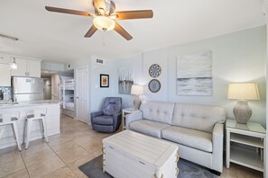 A modern living room with a sofa, armchair, and ottoman, a dining area with a kitchen in the background, and a ceiling fan. - Royal Palms 1008 by Liquid Life (Gulf Shores)
