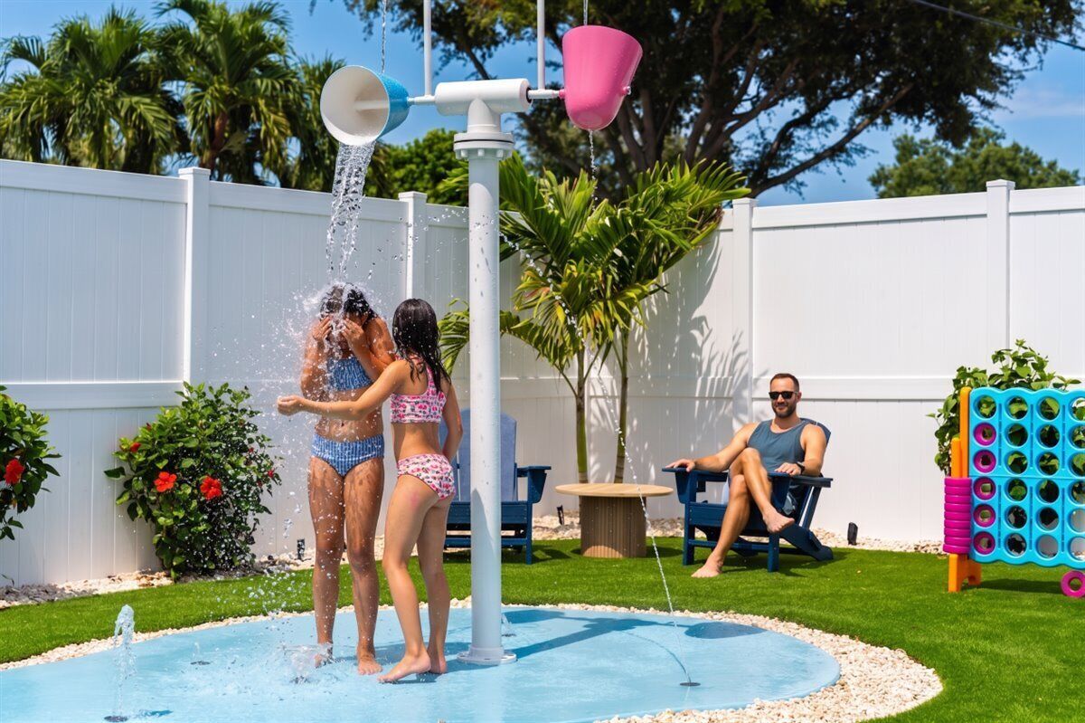 Una piscina con fuente de agua, rodeada por una valla blanca, palmeras y una silla azul.