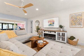 A modern living room with a grey sofa, wooden coffee table, and a fireplace. A ceiling fan and a framed artwork are visible. - Resort-style 5BR/3BA home, 8 min to Anna Maria Island. Private heated pool, splash pad, home theater, arcade, pickleball court, sauna & hot tub — sleeps 18 with a boat-themed bunk room kids love. Everything your group needs, right at home. (Bradenton)