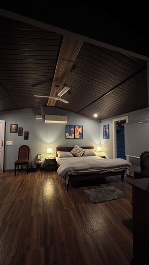 Une chambre avec un plafond en bois, un lit avec une tête de lit, des tables de chevet avec des lampes, une chaise et des œuvres d’art accrochées aux murs.
