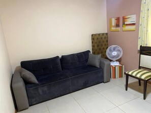 Uma sala de estar com um sofá escuro, uma mesa pequena com um ventilador, uma cadeira com almofada listrada e duas pinturas penduradas na parede.