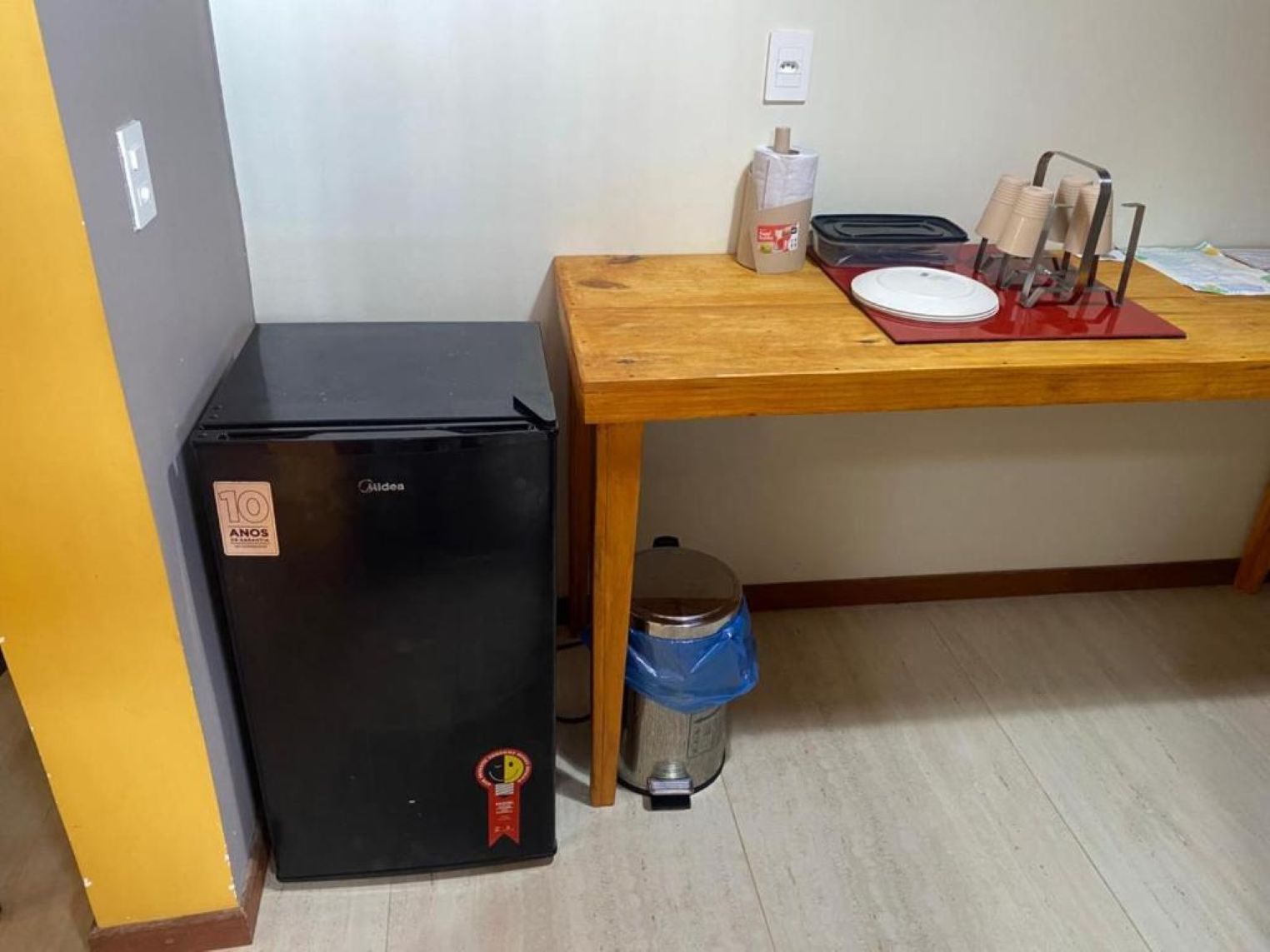 Un refrigerador compacto negro, una mesa de madera con un mantel individual rojo, un cesto de basura y un enchufe de pared.
