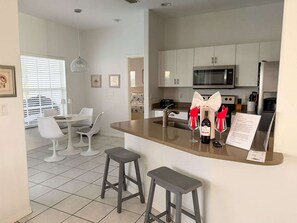 Cocina moderna con armarios blancos, microondas y refrigerador de acero inoxidable. Hay una zona de comedor con mesa y sillas blancas, y una isla de cocina con taburetes.