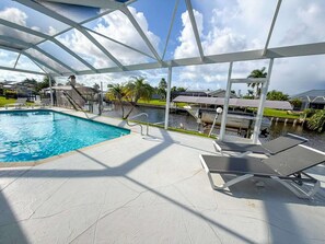 A swimming pool with lounge chairs, a covered area, and a view of a residential area with houses and palm trees. - Villa Dolphin – Prime Location, Southern Exposure, Pool & Gulf Access (Cape Coral)