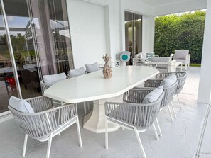 A modern outdoor dining area with a white table surrounded by wicker chairs, a decorative pineapple centerpiece, and a view of greenery through large windows. - Villa Dolphin – Prime Location, Southern Exposure, Pool & Gulf Access (Cape Coral)