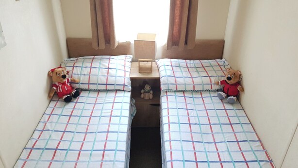 A twin room with two beds, each with a teddy bear, a window with curtains, and a small wooden nightstand. - 10 Berth Caravan Butlins Skegness Pet Friendly (Skegness)