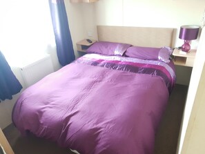 A single bed with a headboard, a bedside table with a lamp, and a window with curtains. - 10 Berth Caravan Butlins Skegness Pet Friendly (Skegness)