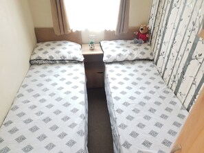 A small, single-room accommodation with two beds, a nightstand, a teddy bear, and a window with curtains. - 10 Berth Caravan Butlins Skegness Pet Friendly (Skegness)