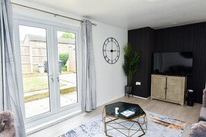 Uma sala de estar com uma mesa de centro de vidro, um relógio na parede e uma televisão em um móvel.