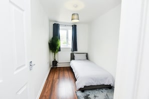 A small, clean room with a bed, a radiator, a window with curtains, and a plant.