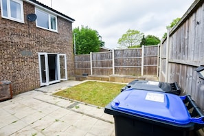 A backyard with a brick house, a wooden fence, and a blue trash bin.
