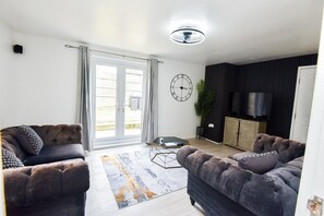 A living room with two sofas, a coffee table, a clock, a television, and a plant. - House Sleeps 7 With Parking (Northampton)