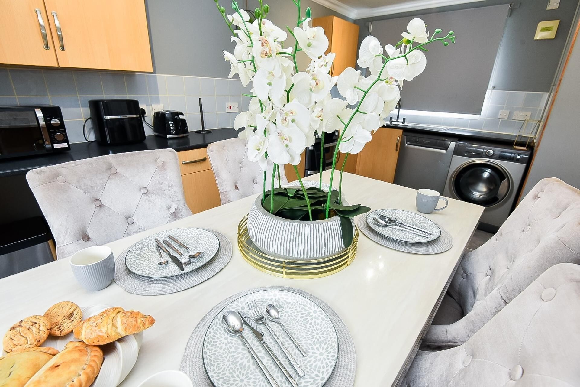 A modern kitchen with a dining table set for two, featuring a vase of white orchids, a coffee machine, and a washing machine in the background.