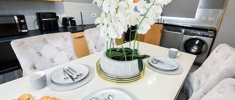 A modern kitchen with a dining table set for two, featuring a vase of white orchids, a coffee machine, and a washing machine in the background.