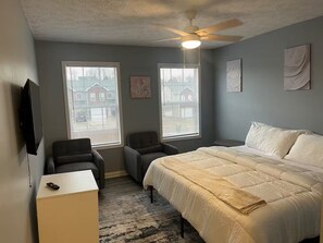 Ein Schlafzimmer mit einem Bett, zwei Stühlen, einem Schreibtisch, einem Deckenventilator und zwei Fenstern mit Jalousien.