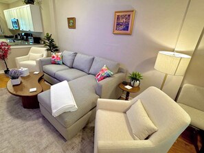 Ein Wohnzimmer mit einem grauen Ecksofa, einem kleinen runden Couchtisch, einem beigen Sessel, einer Stehlampe und einer Küche im Hintergrund.