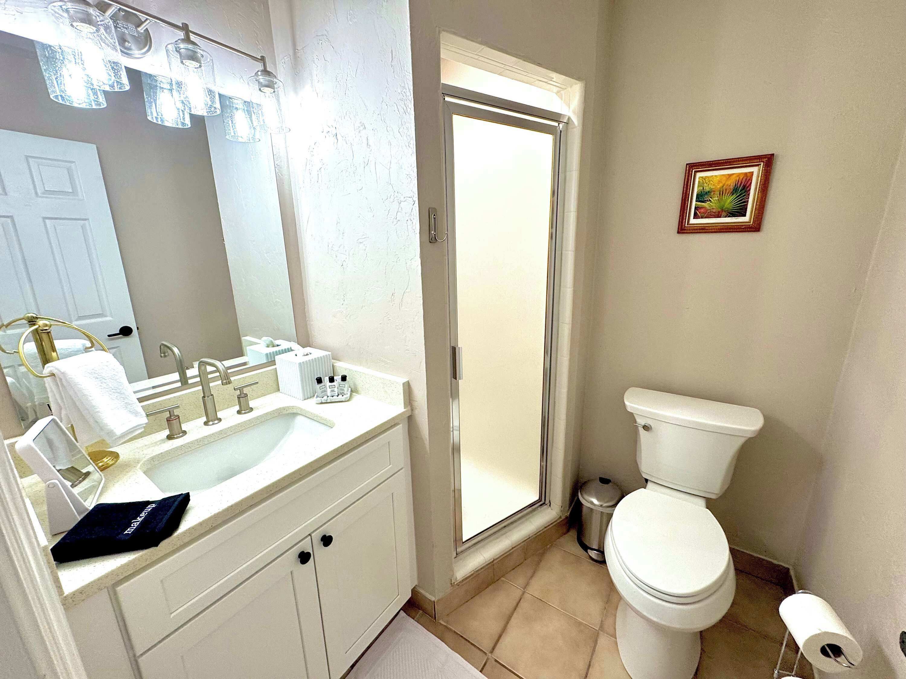 A bathroom with a white sink, mirror, and toilet. There is a towel rack with a towel, a trash can, and a picture on the wall.