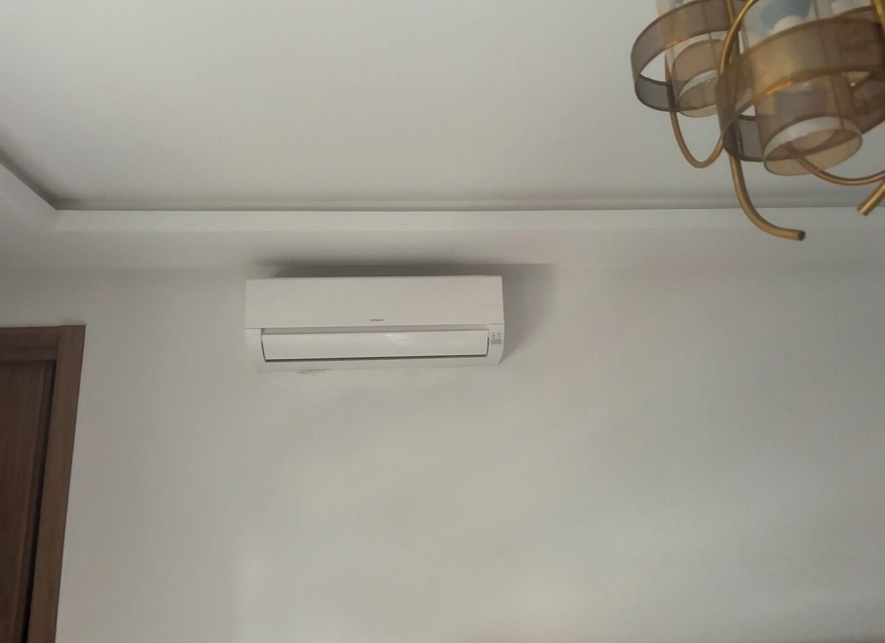 Un appareil de climatisation blanc est fixé au mur dans une pièce dotée d’une porte en bois et d’un luminaire au plafond.