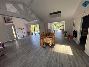 A spacious room with a dining table and chairs, a large window, and a Yamaha sign on the wall. - Forest Cabin on 11 Hectares (Le Bosc du Theil)