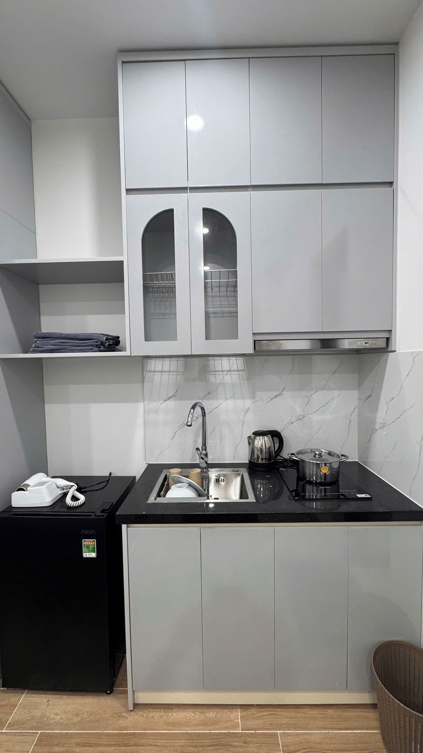 Una cocina compacta con armarios grises, una encimera negra, un fregadero, un hervidor y un refrigerador pequeño.