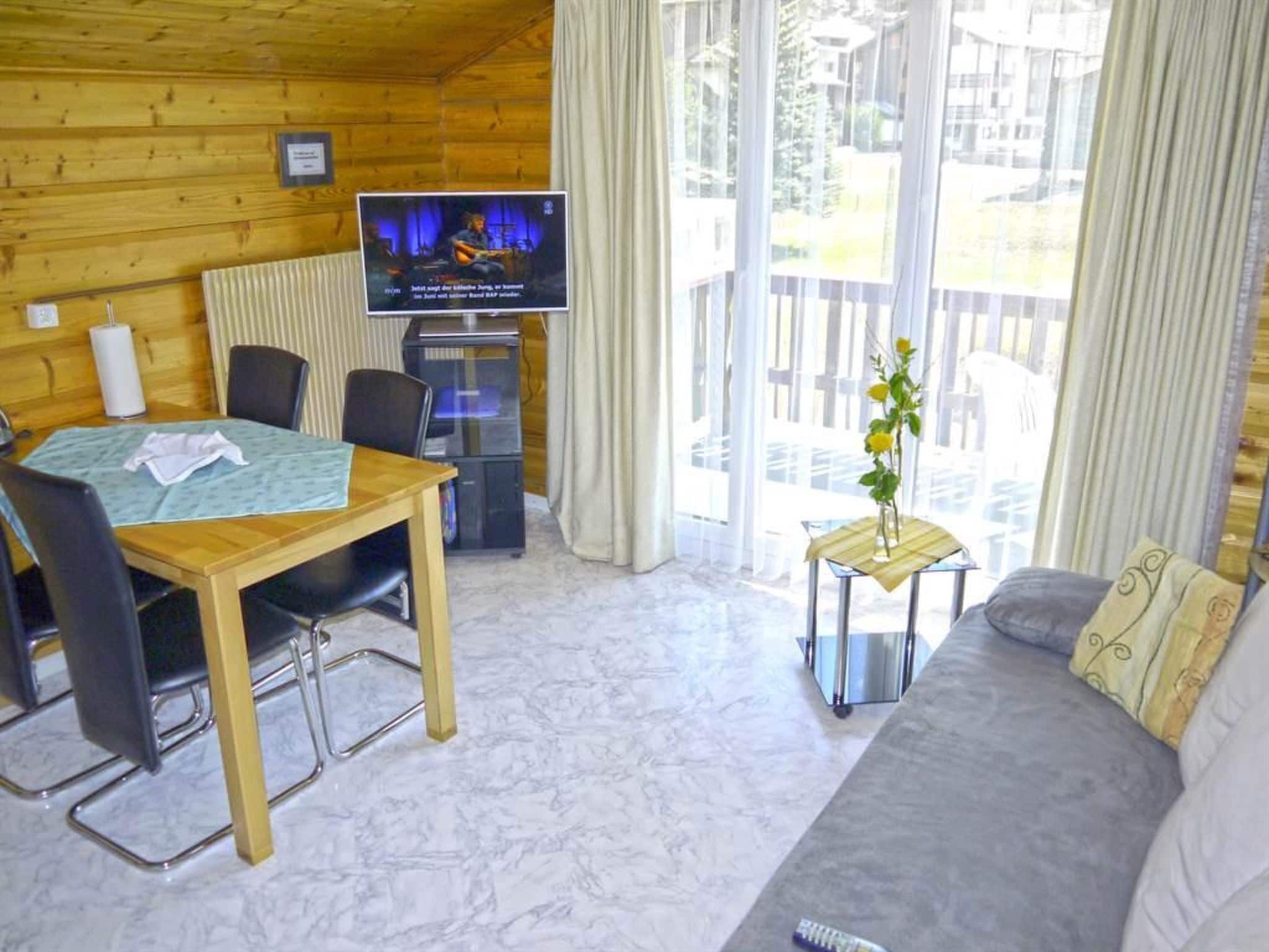 Interior de madera con una mesa de comedor, sillas, un sofá, un televisor y un balcón con cortinas.