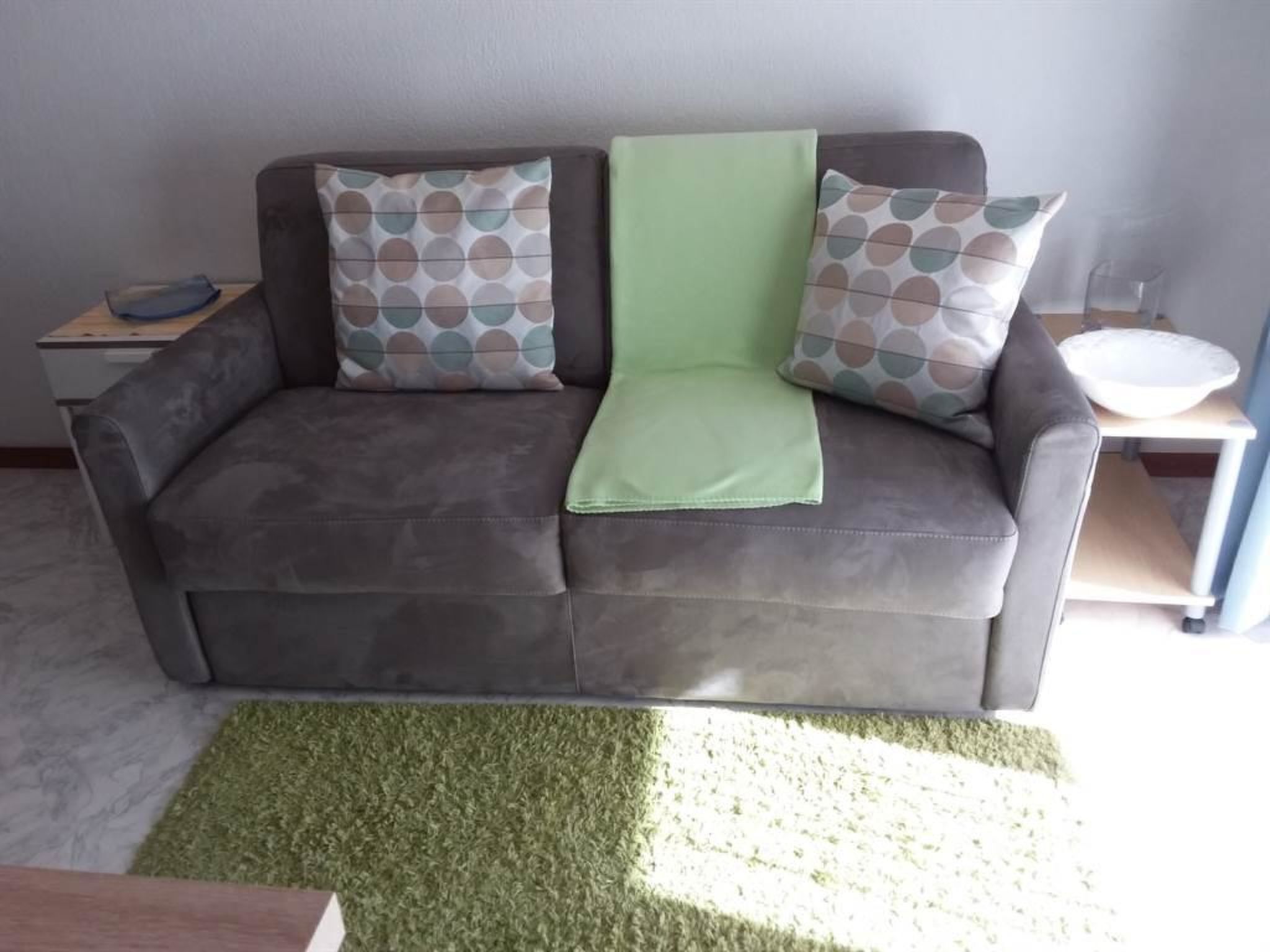 Un sofá gris con cojines estampados, un plaid verde y una alfombra verde en una habitación con pared blanca y una mesita con un bol.
