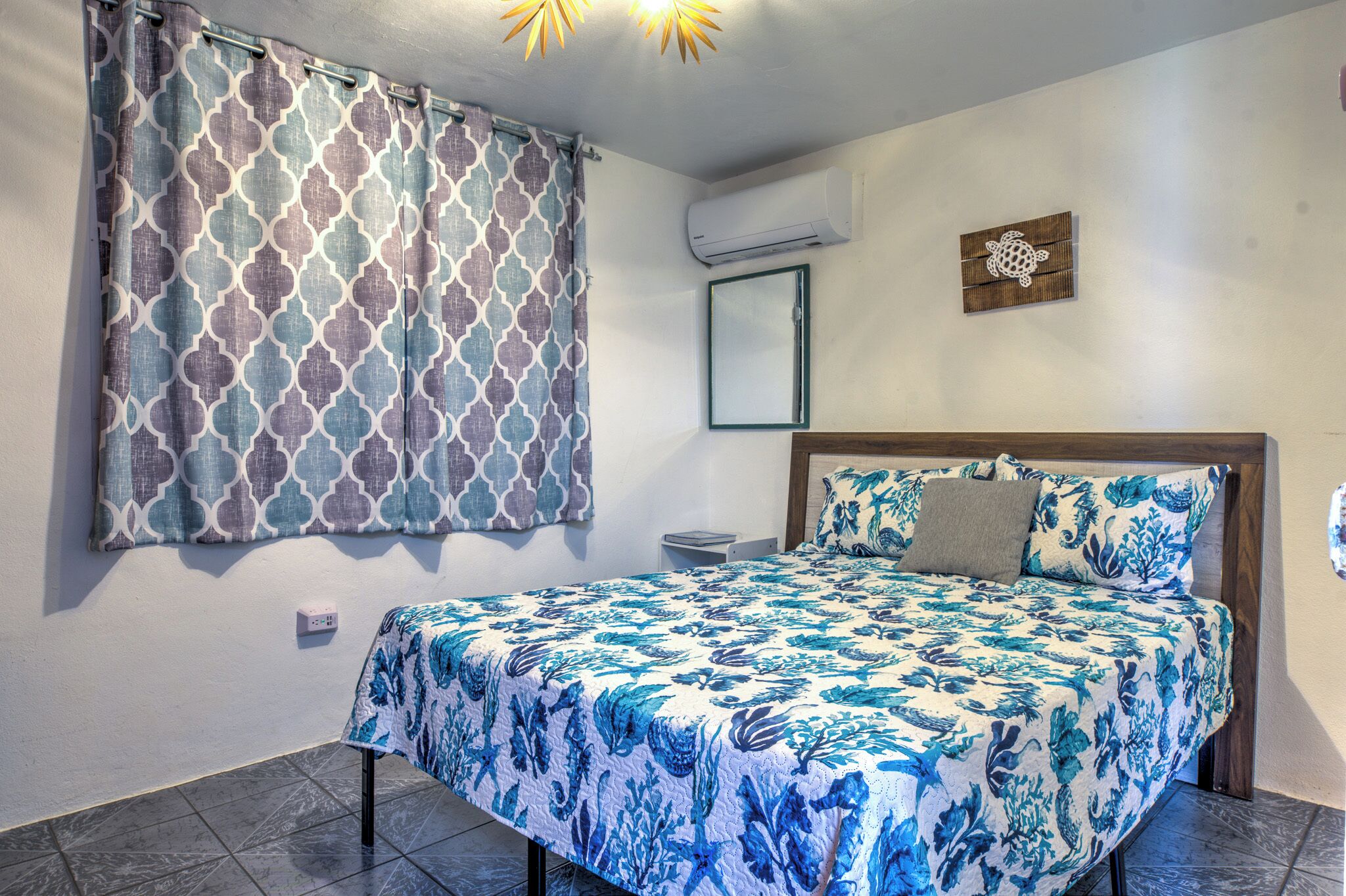 Un dormitorio con cama, cortinas estampadas, aire acondicionado, una mesita pequeña y un espejo instalado en la pared.
