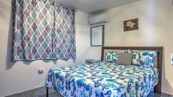 Un dormitorio con cama, cortinas estampadas, aire acondicionado, una mesita pequeña y un espejo instalado en la pared.