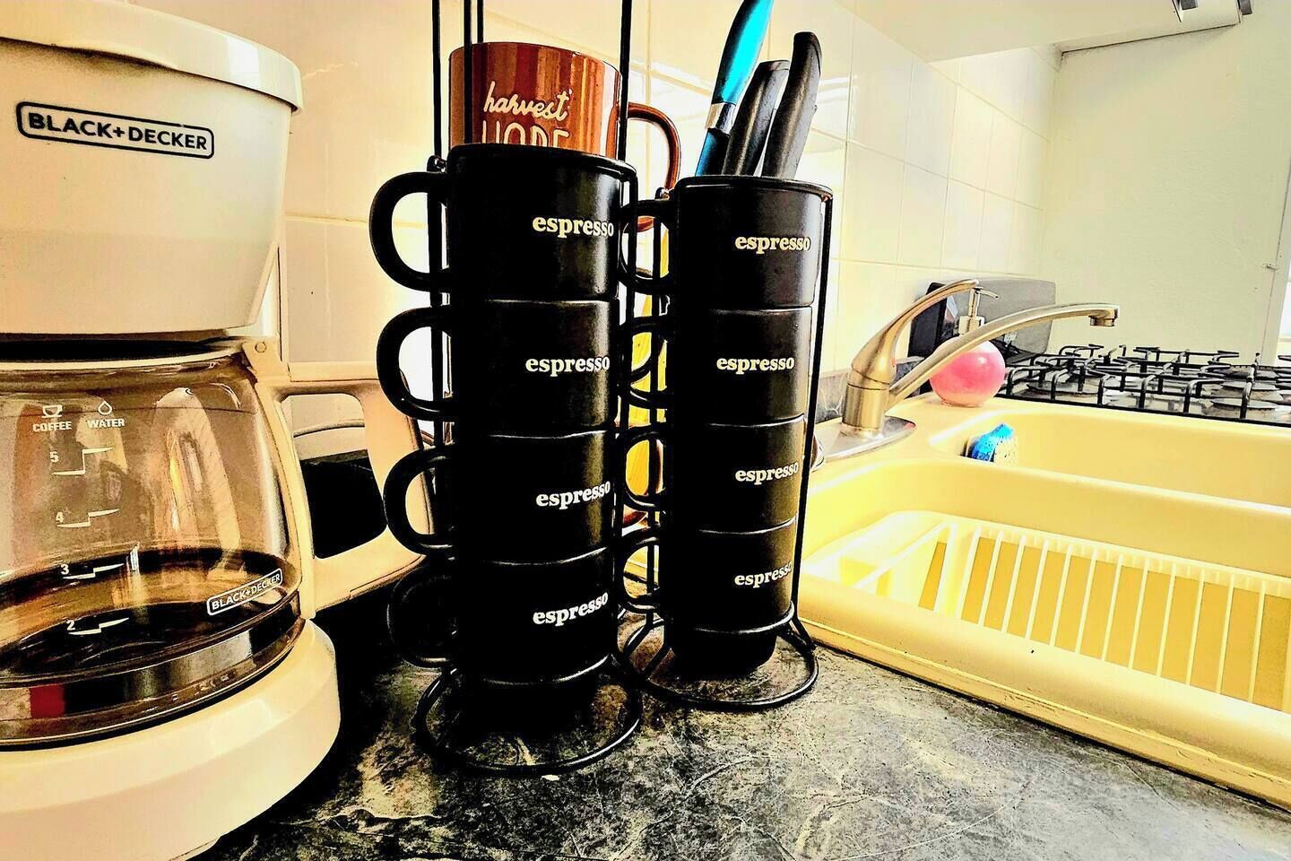 En köksbänk med en kaffebryggare, en hög med espressokoppar och en gul diskho.