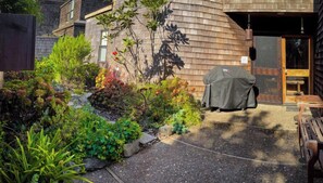 Ein überdachter Außenbereich mit Grill, umgeben von einem Garten mit verschiedenen Pflanzen und Sträuchern.