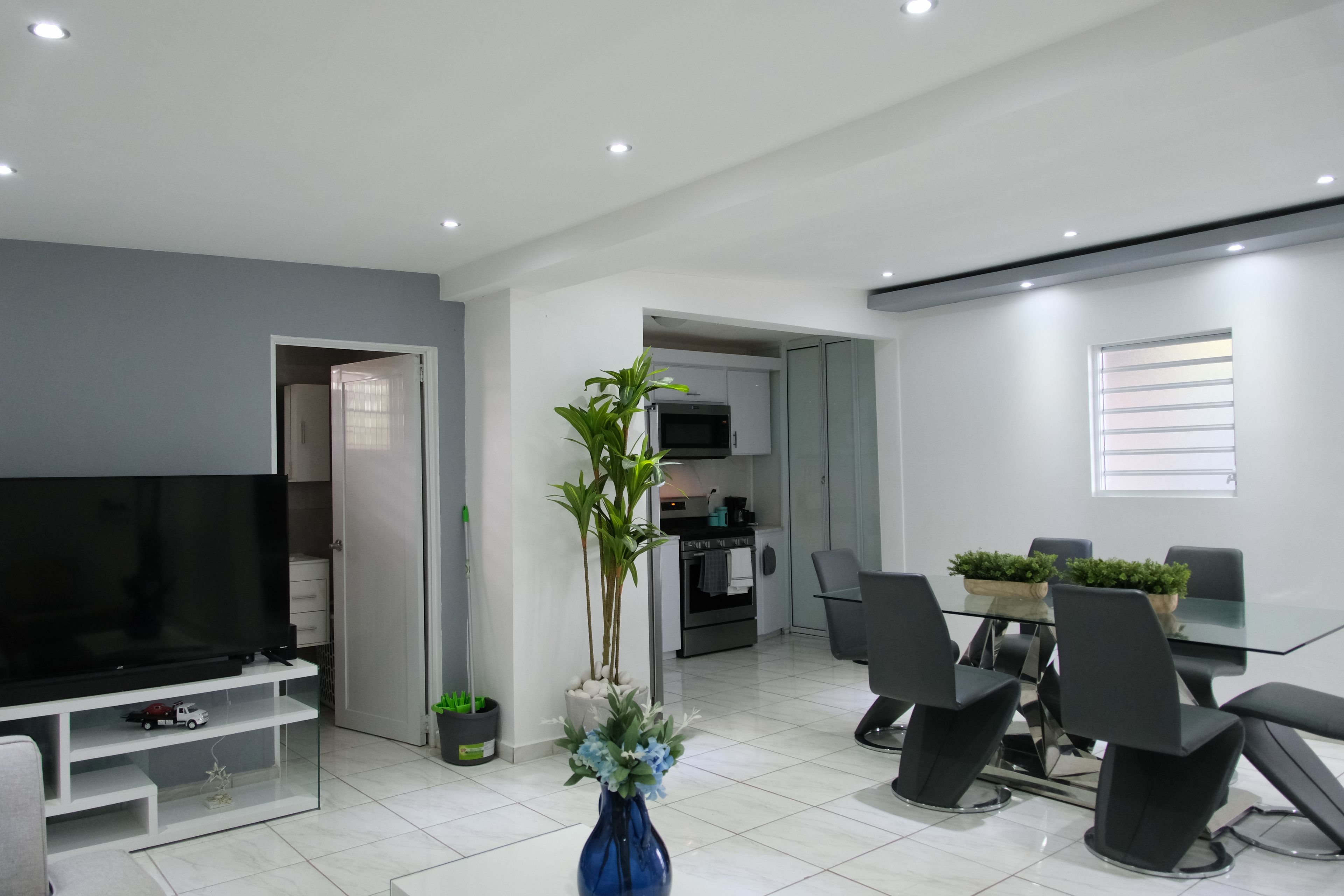 Een moderne woonkamer met een flatscreen televisie, een eethoek met een glazen tafel, en een keuken op de achtergrond.