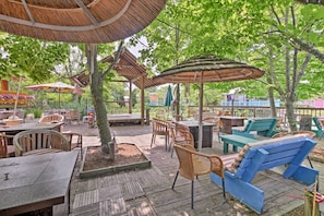 An outdoor patio with wooden tables and chairs, a blue lounge chair, and a thatched umbrella. - Middle Bass Island Escape: Resort Pool & Bar! (Put-in-Bay)