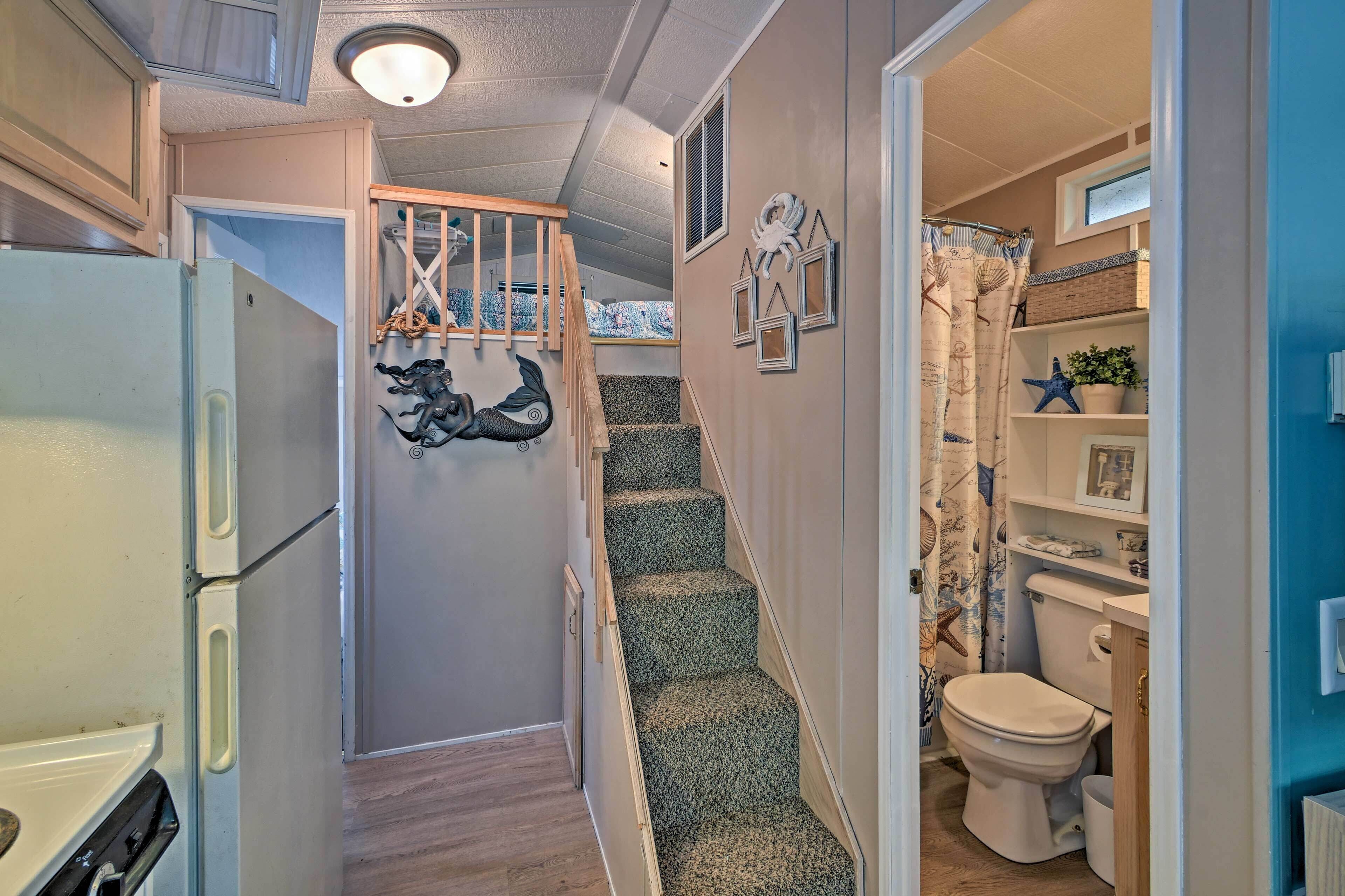 A narrow hallway with a wooden staircase leading to a bedroom. A refrigerator is on the left, and a bathroom with a toilet and shower curtain is on the right.