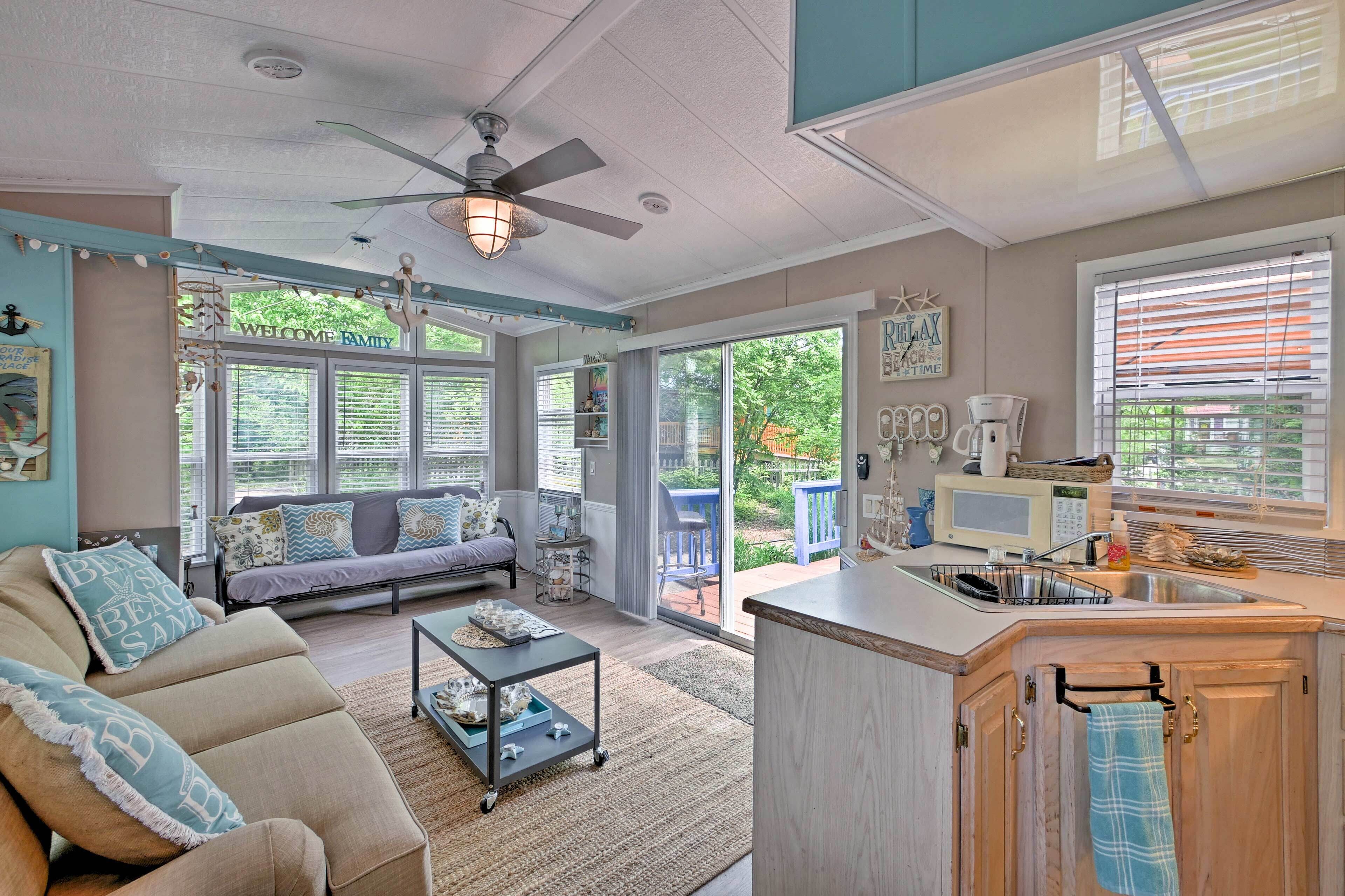 A cozy living room with a sofa, coffee table, and a ceiling fan. There is a kitchen area with a sink and a window.