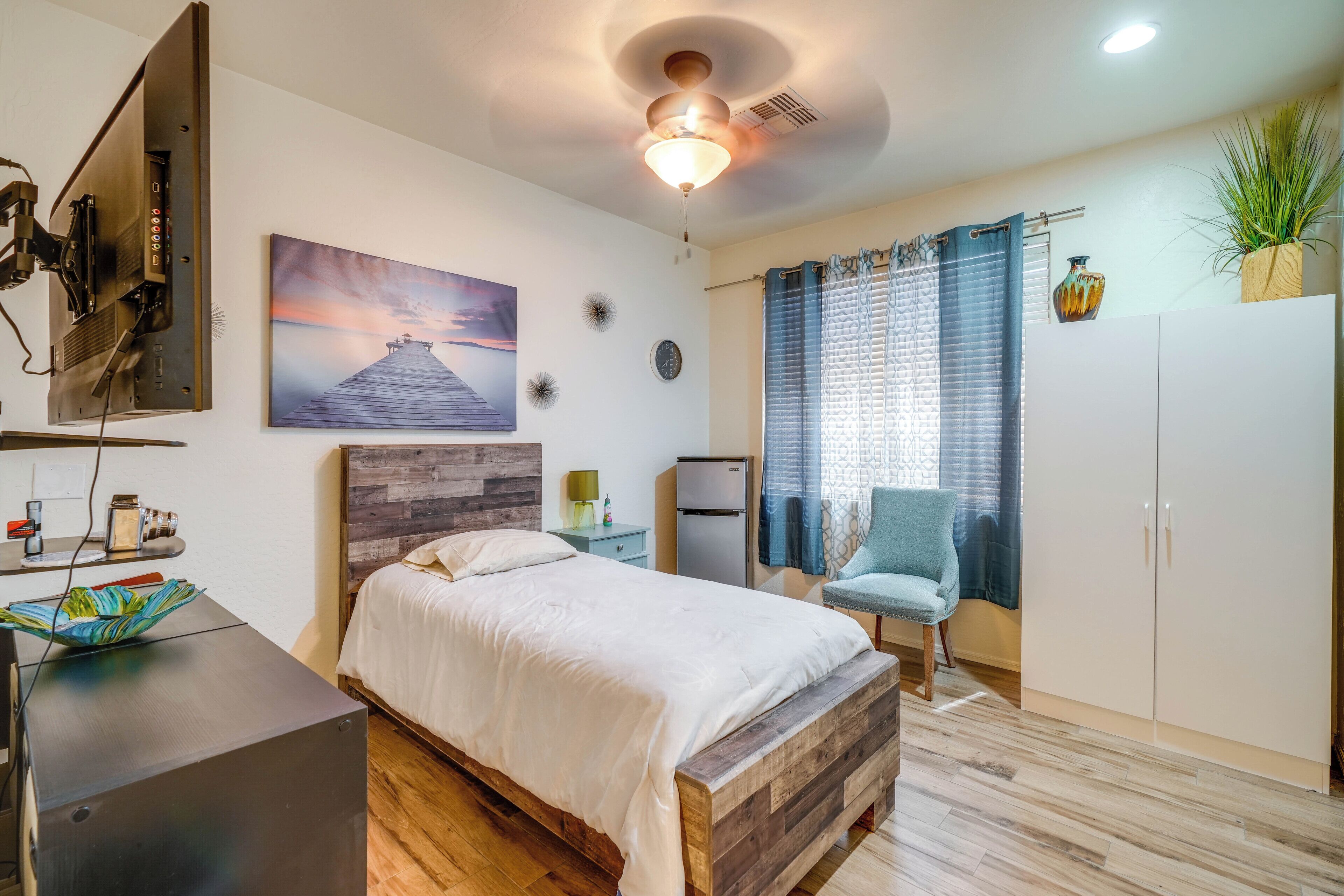 A bedroom with a bed, a desk, a chair, a wardrobe, a plant, and a painting on the wall.