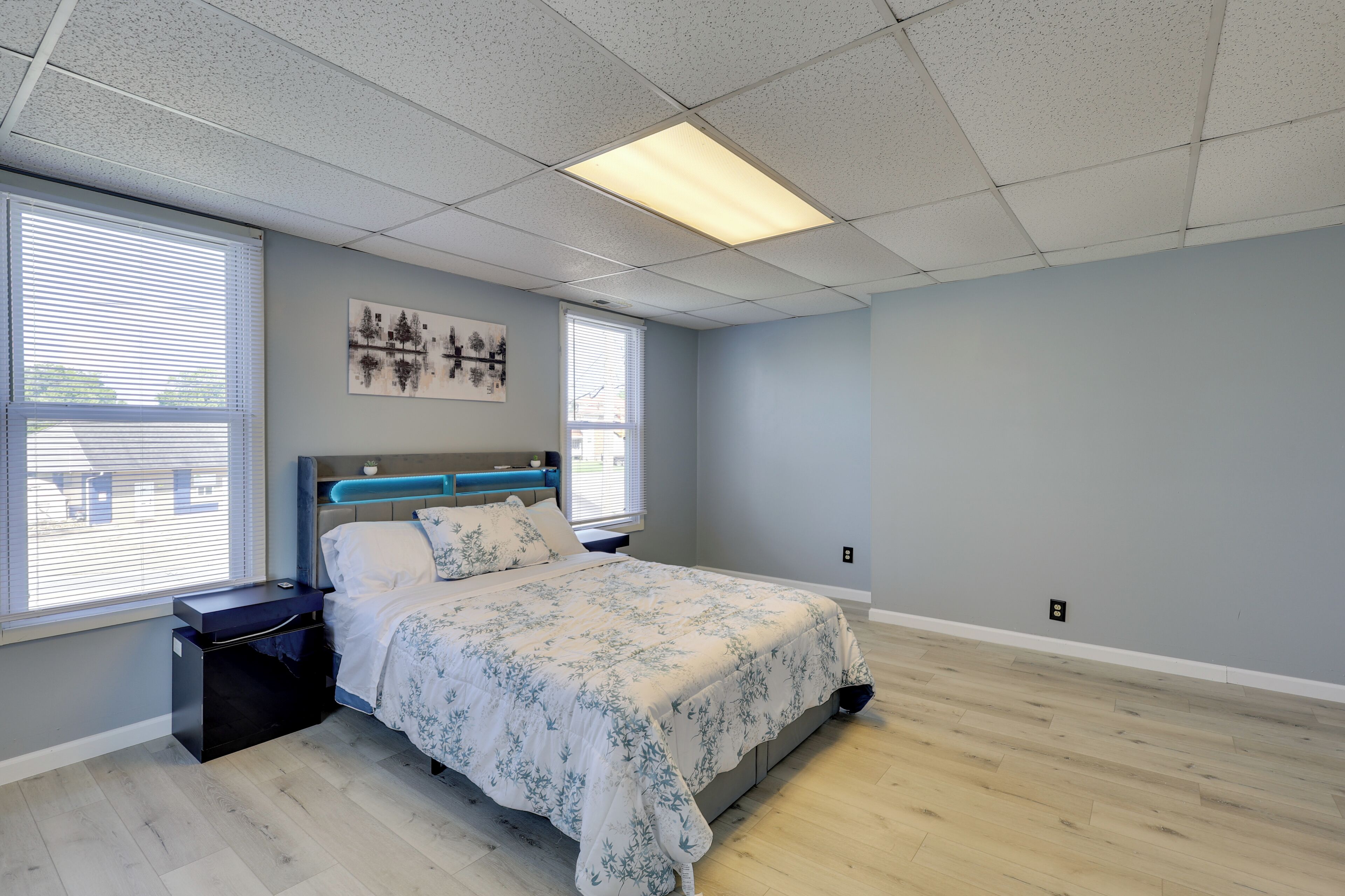 A bedroom with a bed, nightstands, a wall-mounted artwork, and a window with blinds.